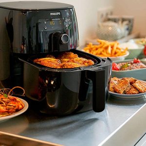 Air fryer cooks chicken; placed on a kitchen counter with plates of fries, prawns, and other foods. 