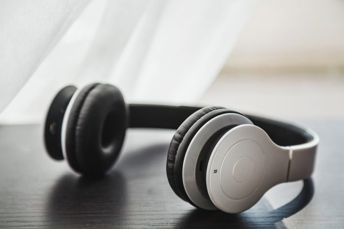 White minimalist wireless headset placed on a dark wooden laminated surface with blurred white curtain crossing a window behind it