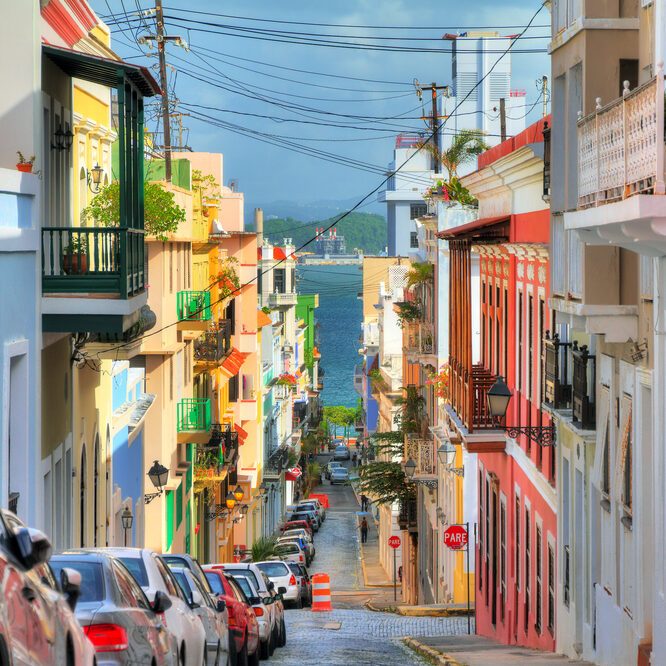 Colorful buildings line a narrow street filled with parked cars, leading down to a distant view of the ocean under a bright blue sky.