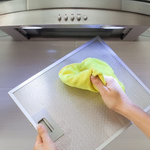 Hands clean a metallic range hood filter with a yellow cloth beneath a kitchen ventilation unit.