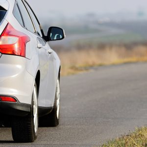A silver car driving on a narrow rural road, surrounded by dry grass and blurred distant fields.