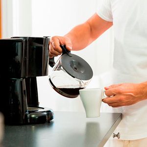 Person pours coffee from a pot into a white mug, standing next to a black coffee maker in a kitchen setting.