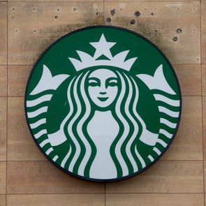 Green circular Starbucks logo; mermaid icon with flowing hair and a crown, displayed against a tan, textured wall background.