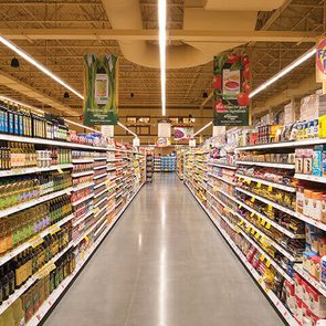 Grocery store aisle displays various food products, neatly organized on both sides, under bright fluorescent lights, with promotional signs overhead highlighting discounts.