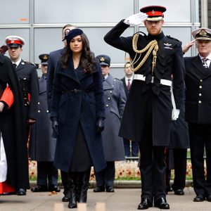 Uniformed individuals stand solemnly, some saluting, during a formal outdoor ceremony with onlookers in military attire, surrounded by poppy wreaths.