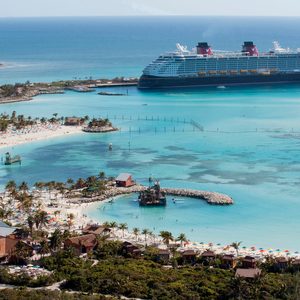A cruise ship docks in a turquoise bay, surrounded by a sandy beach with colorful umbrellas and lush greenery.