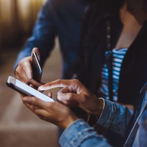 Two people using smartphones in a dimly lit area, focusing on screens with fingers interacting.