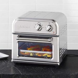 A stainless steel countertop toaster oven is baking vegetables, set against a tiled white wall in a kitchen.