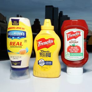 Three condiment bottles stand on a reflective white surface; mayonnaise, mustard, and ketchup with a city skyline in the background.