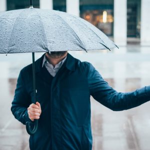 Umbrella shelters person, who gestures with outstretched hand, in a rainy urban setting.