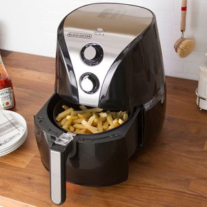 Air fryer holds cooked fries on a wooden countertop, surrounded by ketchup, stacked plates with napkins, and kitchen items.