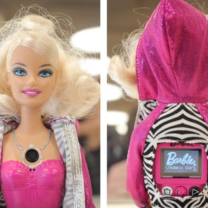 Doll wears pink and silver outfit, displaying a camera on chest. Back reveals 