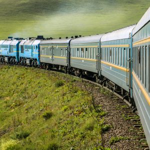 Train traveling along a curved, grassy terrain with bright blue engines and passenger cars, surrounded by open fields.