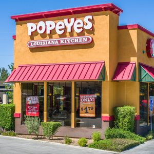 Popeyes building featuring red awnings and promotional posters, located in a parking lot with shrubs and a Starbucks coffee shop in the background.