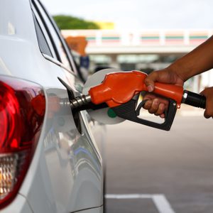 A person refuels a white car with an orange gas nozzle at a filling station, the background visibly blurred.
