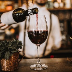 Wine bottle pours red wine into glass; bar setting with potted plant and skewers nearby.