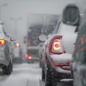 Cars are stopped on a snow-covered road, brake lights glowing red amidst falling snow, in a gray, overcast atmosphere.