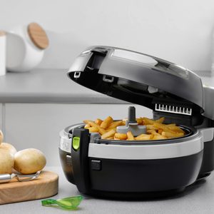 A black air fryer cooks fries on a kitchen countertop, surrounded by fresh potatoes, a peeler, and a jar with a wooden lid.