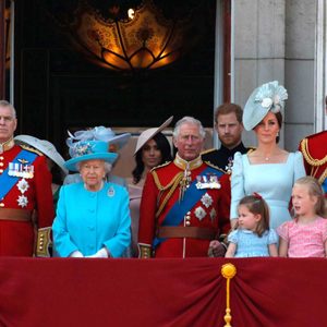 Adults and children stand on a balcony, dressed in formal attire and uniforms, against a backdrop of ornate architecture.