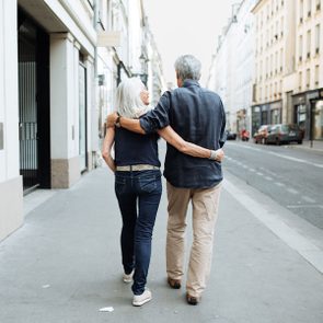 A couple walks arm-in-arm along a quiet, urban street, flanked by tall buildings and parked cars.