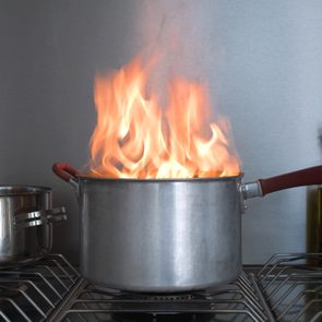 Pot engulfed in large flames sits on a stove; nearby bottles and pepper grinder in a kitchen setting.
