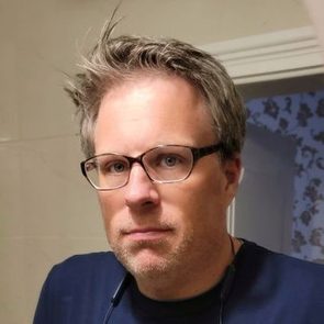 A person with glasses looks directly ahead in a bathroom, with light-colored walls and a slightly opened door in the background.