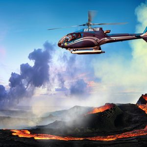 Helicopter flies over an erupting volcano, with lava flowing and smoke billowing in a vibrant sunset sky. Text on helicopter: 