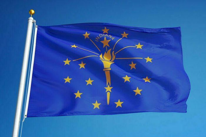 A blue flag with a torch and stars waves on a flagpole against a clear sky, displaying "INDIANA" above the torch.