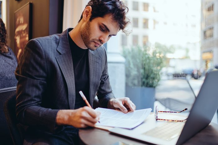 Young handsome caucasian businessman in trendy suit writing information on financial papers for banking on laptop computer, serious confident male doing paperwork remotely indoors in coffee shop
