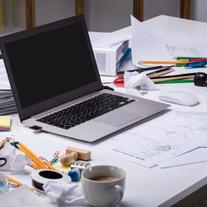 Laptop sits idle on a cluttered desk, surrounded by scattered papers, stationery, folders, and a coffee cup in an office environment.