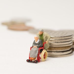 Miniature figures: a person pushes another in a wheelchair beside stacks of coins, symbolizing financial planning and elderly care.