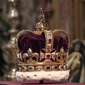 A jeweled crown rests on a cushion, surrounded by ornate decor in a regal setting.