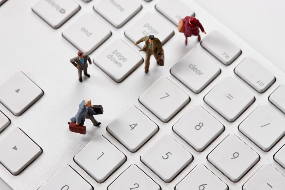 Miniature figures walking on a computer keyboard, positioned between large keys, suggesting an imaginative blend of business and technology.