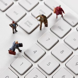 Miniature figures walking on a computer keyboard, positioned between large keys, suggesting an imaginative blend of business and technology.