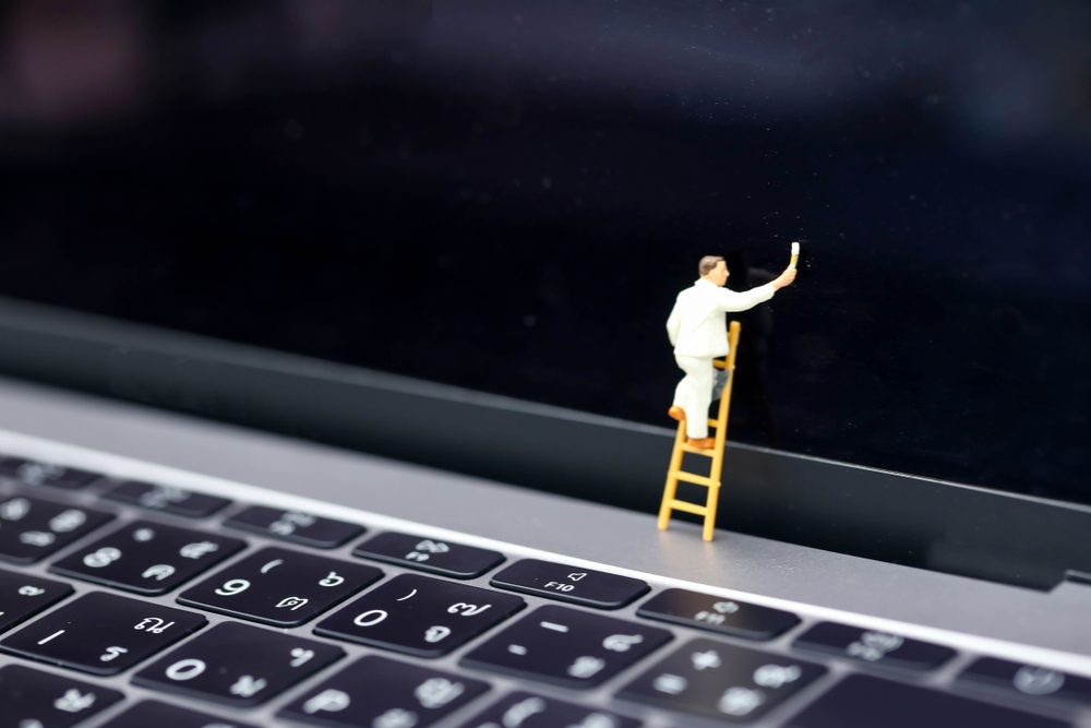Miniature figure paints on a laptop screen, standing on a small ladder above the keyboard, creating a surreal scene.