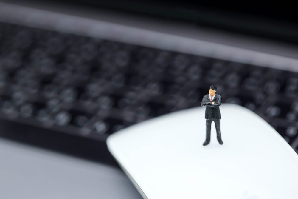 A small toy figure stands on a computer mouse, with a blurred keyboard in the background.