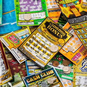 Various scratch-off lottery tickets overlapped under coins, displaying colorful designs and potential prizes in a mixed stack.