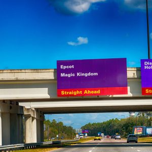 Highway signs direct drivers; one lists Epcot and Magic Kingdom, and another shows Disney's Hollywood Studios, all against a clear blue sky.