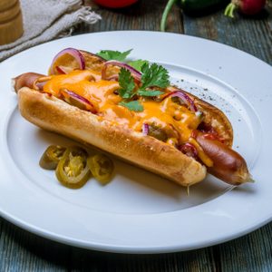 A hot dog topped with cheese, red onions, cilantro, sits on a white plate with jalapeÃ±os, surrounded by peppers and napkins on a wooden table.