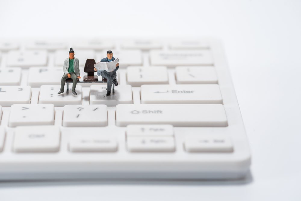 Miniature figures sit on a white keyboard, one reading a newspaper, the other relaxing, creating a surreal office scene on the keys.