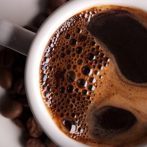 A steaming cup of black coffee, bubbles on surface, rests on a white saucer surrounded by scattered coffee beans.