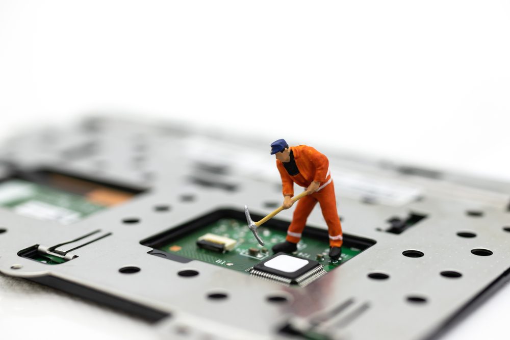 A tiny worker figurine digs with a pickaxe on a computer circuit board, creating a humorous scene of technology and manual labor.