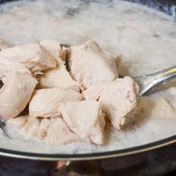 Chunks of chicken are being boiled in frothy water on a stovetop, with a slotted spoon lifting them from the pot.