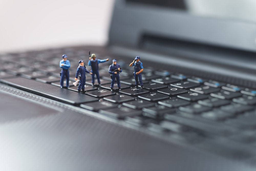 Miniature figures stand on a laptop keyboard, appearing as workers inspecting keys. The background is blurred, emphasizing the scale contrast.
