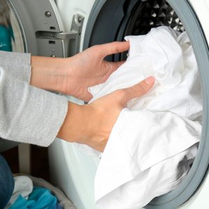 Hands place white clothes into a front-loading washing machine; assorted laundry items are visible nearby.