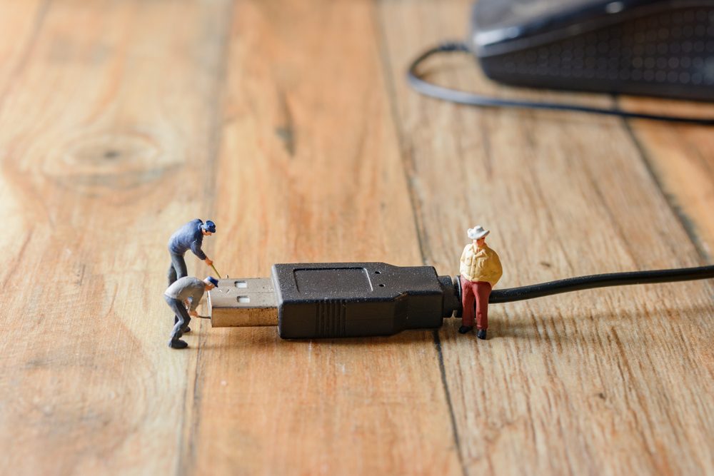 Tiny figurines examine a giant USB plug on a wooden surface, appearing to work or inspect it.