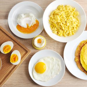 Various egg dishes, including poached, scrambled, boiled, fried, and an omelette, are displayed on white plates and a wooden surface.