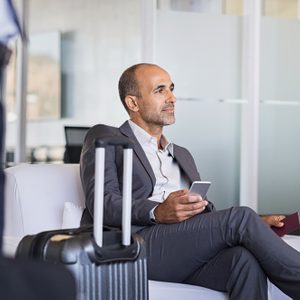 Man sits holding phone and passport, next to suitcase, in a modern office-like waiting area, looking thoughtful.
