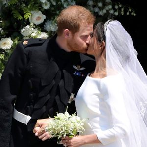 A couple is kissing, wearing wedding attire, surrounded by a backdrop of lush white roses and greenery.