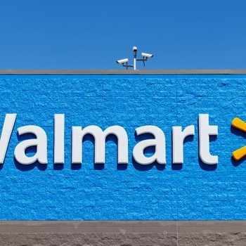 "Walmart" sign displayed on a blue brick wall with a security camera above; clear sky background.